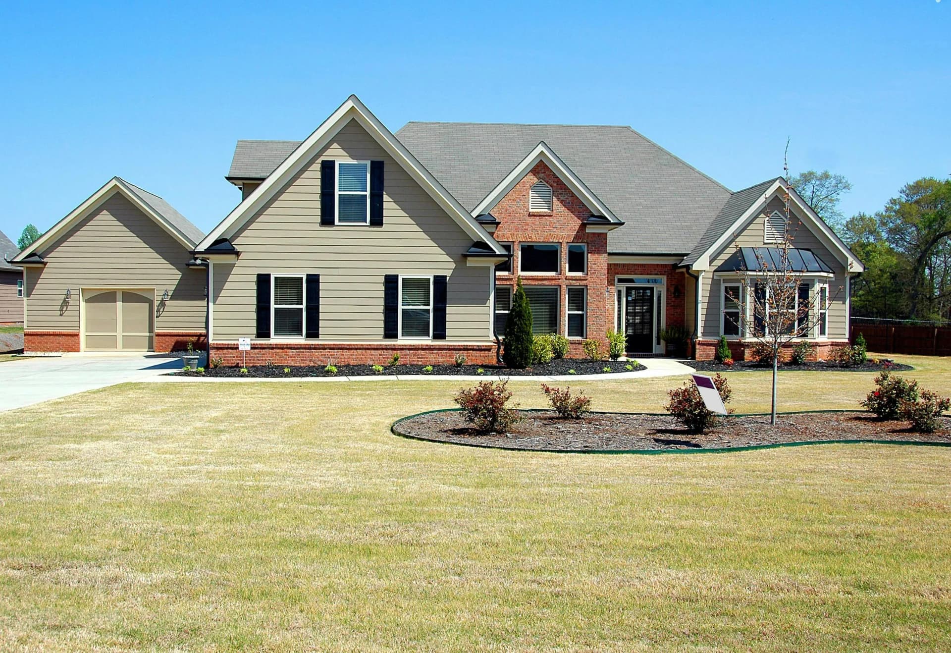 A large, modern detached house with beige siding, brick accents, multiple gables, and a grey shingle roof, set against a bright blue sky with a well-maintained lawn and landscaped garden beds in the foreground.