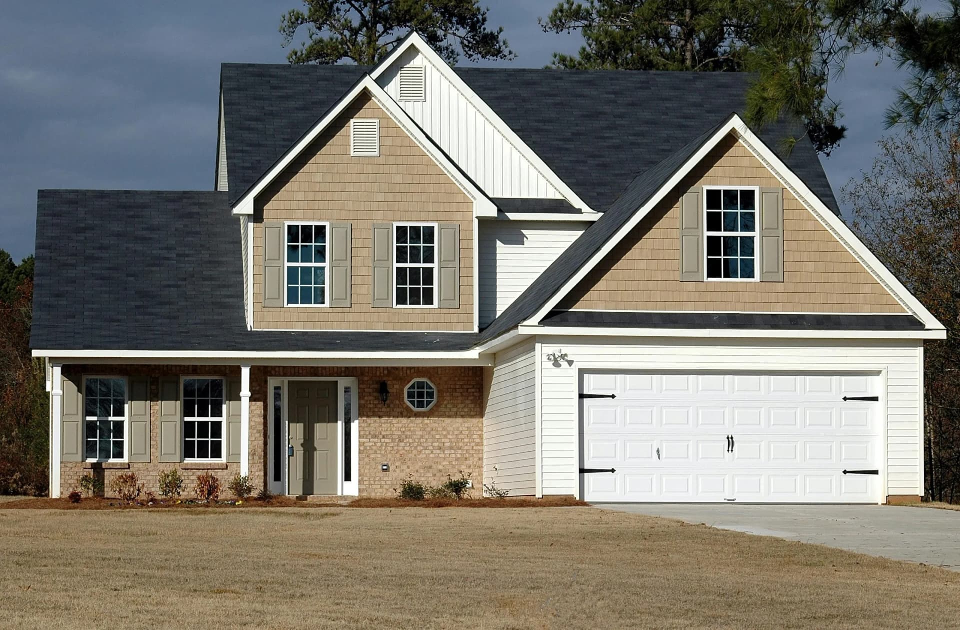 A modern two-story suburban house with a dark grey roof, light brown siding, and a white garage door, set against a cloudy sky.