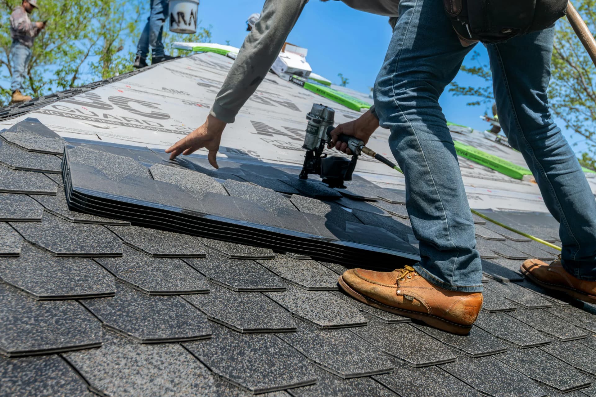 A roofer installing dark shingles on a new roof using a nail gun, with underlayment visible.